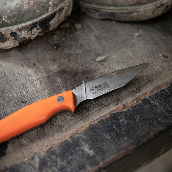 A Casström Swedish Field Dresse knife with orange g10 handle orthogonal view, with gumboots in the background.